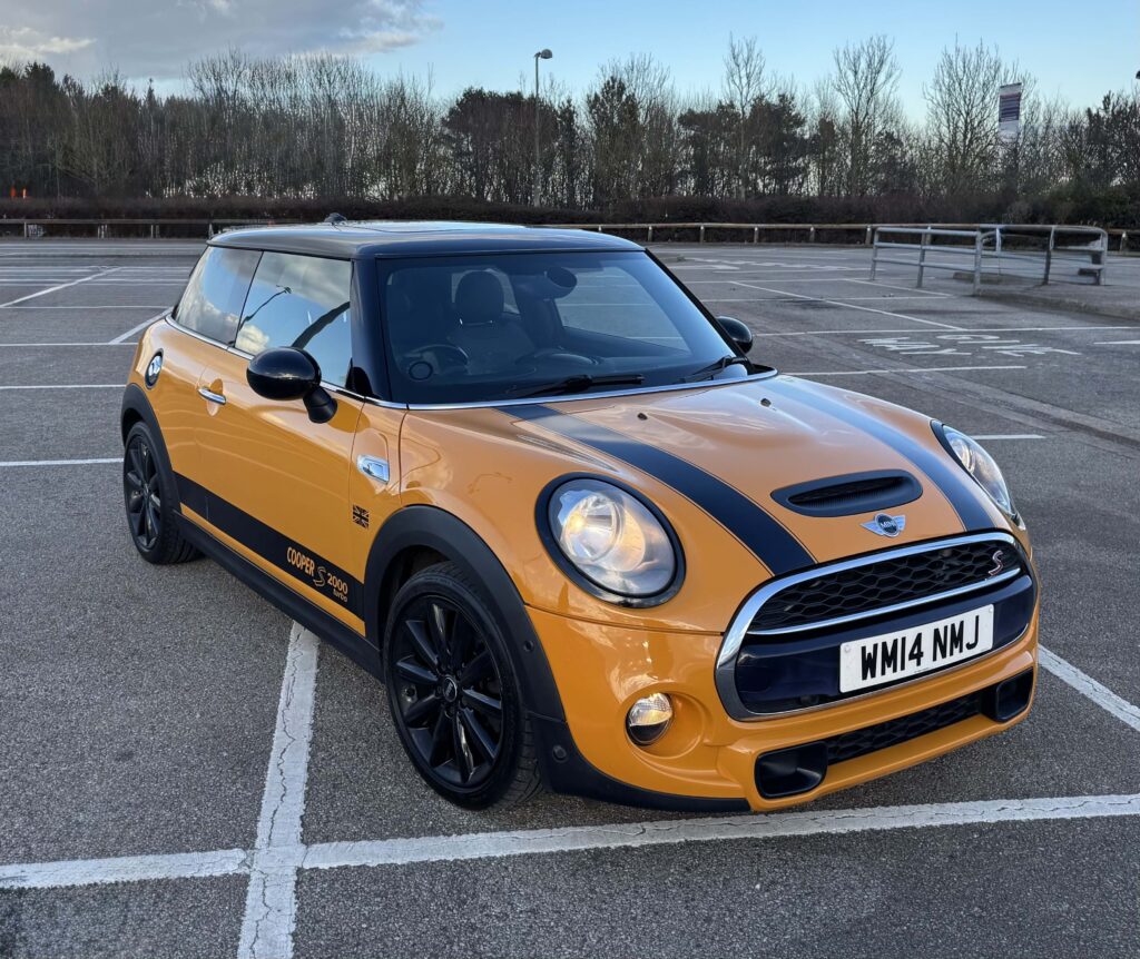 A bright orange Mini Cooper S with black racing stripes and black wheels is parked in an empty outdoor lot. This stylish mini car’s headlights are on, and trees line the background under a clear sky. The license plate reads "WM14 MNJ.