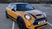 A bright orange Mini Cooper S with black racing stripes and black wheels is parked in an empty outdoor lot. This stylish mini car’s headlights are on, and trees line the background under a clear sky. The license plate reads "WM14 MNJ.