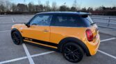A yellow Mini Cooper S with a black roof, black wheels, and racing stripes is parked diagonally in an empty lot. Perfect for any mini trader or enthusiast, this mini car stands out against trees and bushes under a partly cloudy blue sky.