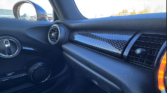 View of a Mini car’s front passenger seat area, featuring a black textured dashboard with a carbon-fiber panel, circular air vents, door handle, and part of the seat. The side mirror and clear sky are visible through the window.