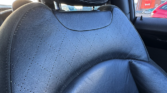 Close-up of a black leather mini car seat with stitched diamond patterns, visible headrest, and sunlight casting shadows. The car window shows other parked vehicles and a red B&Q store in the background.