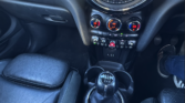 Interior of a MINI car showing the dashboard, manual gear stick, climate controls, and infotainment screen with parking assist. Black leather seats and the driver’s right foot in a sneaker are partially visible. Perfect for MINI traders.