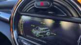 A mini car dashboard display shows a tire pressure monitor screen with all four tires at 2.3 bar. The circular display is edged by an orange LED ring, set within a textured carbon fiber panel and topped by a hazard light button. Perfect for any MINI trader.