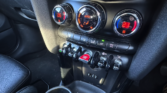 Close-up of a MINI car’s center console featuring three circular climate control dials with digital displays, toggle switches below for various functions, cup holders at the bottom, and black leather seats partially visible on the left.