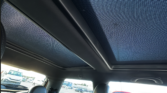 View from inside a mini car showing a panoramic sunroof with mesh covering, two front seats, and parts of the parking lot outside—a red car is visible through the window on the right, with other vehicles seen in the distance.