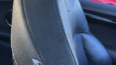 Close-up of a black leather MINI car seat with the MINI logo stitched near the edge. Outside the window, a red mini and several other vehicles are visible under a partly cloudy sky—perfect for any Mini trader's showroom.