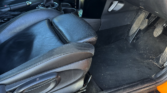 Interior view of a MINI Cooper S mini car, focusing on the black leather driver's seat, floor mat, pedals, and lower dashboard. The groove-marked door sill plate shows "MINI COOPER S" in silver and red. The car door is open.