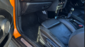 The photo shows the interior of an orange Mini Cooper S, focusing on the passenger seat, dashboard, and gearshift. The car door is open, revealing black leather seats and a sporty mini car dashboard with round vents.