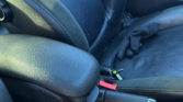 A close-up view of a mini car’s interior focuses on the black leather seats. A black glove rests on the passenger seat, while the center console, handbrake with a red release button, car keys, and circular control knob are visible.