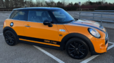 A bright orange mini car with a black roof, black wheels, and "COOPER S 2000" plus a UK flag decal on the side, parked in an empty outdoor lot with trees and a cloudy sky in the background.