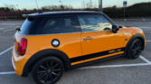 A bright orange Mini Cooper S with a black roof and mirrors is parked in an empty lot. This stylish mini car features black wheels, a "Cooper S 2000" side stripe, and a chrome fuel cap. The sky is partly cloudy with shrubbery in the background.