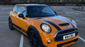 A bright orange Mini Cooper S with black stripes on the hood is parked in an empty lot. This sleek mini car features black rims, tinted windows, and a black roof. Bare trees and an overcast sky create a moody backdrop for this Mini Trader find.