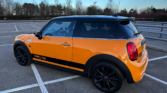A vibrant orange Mini Cooper S mini car with a black roof and black wheels is parked diagonally in an empty lot. “Cooper S 2000” decals stand out on the side, framed by leafless trees and a clear blue sky.