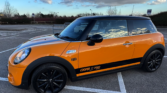 A bright orange Mini Cooper S with black stripes, black wheels, and black mirror caps is parked in a nearly empty outdoor lot. “COOPER S 2000” and a Union Jack badge mark this stylish mini car. The sky is cloudy with blue patches.