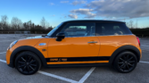 A yellow Mini Cooper with black roof and wheels is parked in an empty lot. This stylish mini car features “COOPER S 2000” decals and a Union Jack flag on the side, set against trees and a cloudy blue sky.