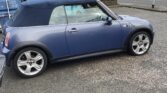 A blue Mini Cooper convertible with a dark soft top is parked on a paved street near a green shipping container and blue bins. Residential houses and leafless trees are visible in the background under a cloudy sky—an ideal find for any mini car enthusiast.