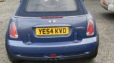 A blue Mini Cooper convertible, listed on Mini Trader, is parked on a residential street and viewed from the rear. The license plate reads "YE54 KVD." Houses, a white garage, and a white van with orange graphics are seen in the overcast background.