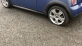 A blue Mini Cooper convertible with a dark blue soft top is parked on asphalt. The rear driver-side tire of this mini car is completely flat, while the others are inflated. A white garage door stands in the background.