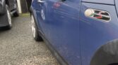A blue Mini Cooper S mini car is parked on a gravel driveway near a garage. The photo shows the driver’s side, including the mirror, badge, and part of the wheel. Another dark vehicle is partially visible beside it under cloudy skies.