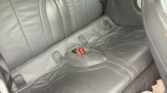 A close-up of the back seat in a mini car, featuring black leather upholstery with three seatbelt buckles, one of which is red. The seat curves upward in the center. The floor has dark carpeting and a cup holder on the right side.
