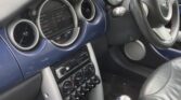 The photo shows the interior of a Mini Cooper car, focusing on the dashboard, steering wheel, manual gear shift, and center console. This stylish mini showcases three round gauges, toggle switches, a radio, blue trim, and leather seats in the foreground.