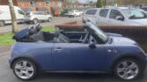 A blue convertible Mini Cooper, listed on Mini Trader, is parked near a brick wall with its roof down, revealing black seats. Other cars and residential houses appear in the background under a cloudy sky.