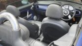 View of a mini convertible car’s black leather interior from above, showing two front seats, a chrome roll bar, circular dials on the dashboard, and an open roof. A brick wall, window, and another parked car are visible in the background.