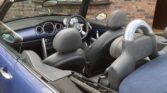 A blue Mini convertible with the roof down is parked in front of a brick house with brown-framed windows. The car’s black and grey interior, steering wheel, headrests, and a silver roll bar behind the seats are visible.