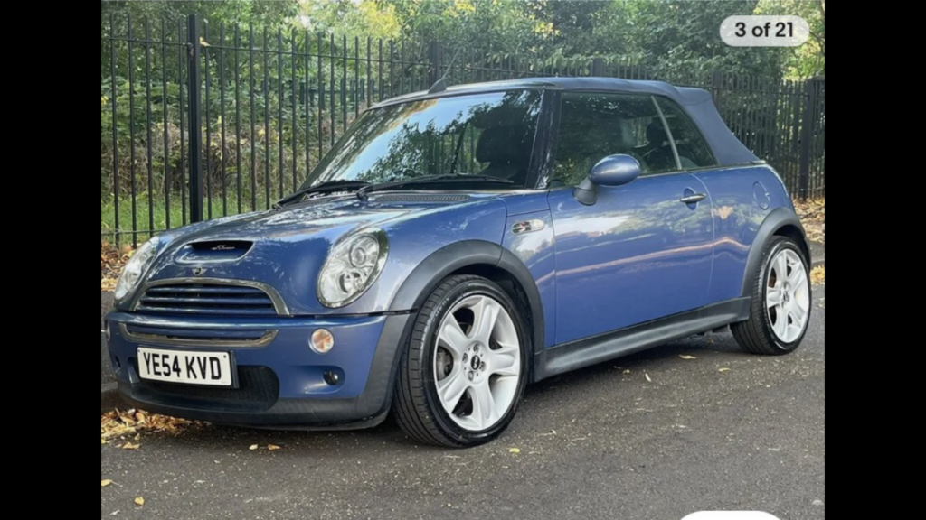 A blue Mini Cooper convertible with a black soft top is parked on a paved road near a black iron fence, surrounded by green trees and scattered dry leaves—a stunning mini car perfect for any Mini Trader. The weather appears clear and sunny.