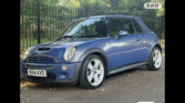 A blue Mini Cooper convertible with a black soft top is parked on a paved road near a black iron fence, surrounded by green trees and scattered dry leaves—a stunning mini car perfect for any Mini Trader. The weather appears clear and sunny.