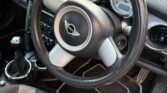 Close-up view of the interior of this iconic mini car, focusing on the steering wheel with the Mini logo, manual gear shift, and dashboard featuring large, round speedometer and dials. The upholstery is two-tone black and grey.