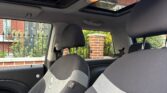 The photo shows the interior front seats of a mini car with a sunroof open, taken from the rear seat. Outside the car, a red brick building with ornate windows, a black iron fence, and some greenery is visible.