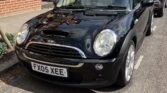 A black Mini Cooper, the classic mini car with license plate "FX05 XEE," is parked on a sunny residential street beside a red-brick house with a small garden and black railings. Another car is parked behind it, with tree shadows falling across the Mini.