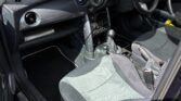 Photo of the interior of a right-hand drive Mini Cooper S mini car. The car door is open, revealing black and grey fabric seats, dashboard with round gauges, manual gear shift, cup holders, and sunlight shining on the seats and floor.