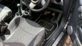 View of a mini car interior from the open driver’s door, showing black and grey seats, steering wheel with Mini logo, circular speedometer and gauges, manual gear stick, metallic foot pedals, and a glimpse of the exterior sidewalk.