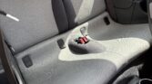 The photo shows the back seat of a mini car with light gray upholstery, black headrests, and a small center hump featuring two red seat belt latches. Sunlight highlights part of the seat, and a yellow UK license plate is visible outside the rear window.