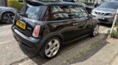 A black Mini Cooper S mini car is parked on a residential street beside the curb. The car has a yellow UK license plate (FV05 XEE) and silver alloy wheels. Other cars and traditional British houses with green hedges are visible in the background.