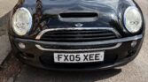 A black mini car is parked on a residential street. The car's front is visible, showing its headlights, grille, and UK license plate "FX05 XEE." Trees, a sidewalk, and other parked cars line the background. The weather appears clear and sunny.