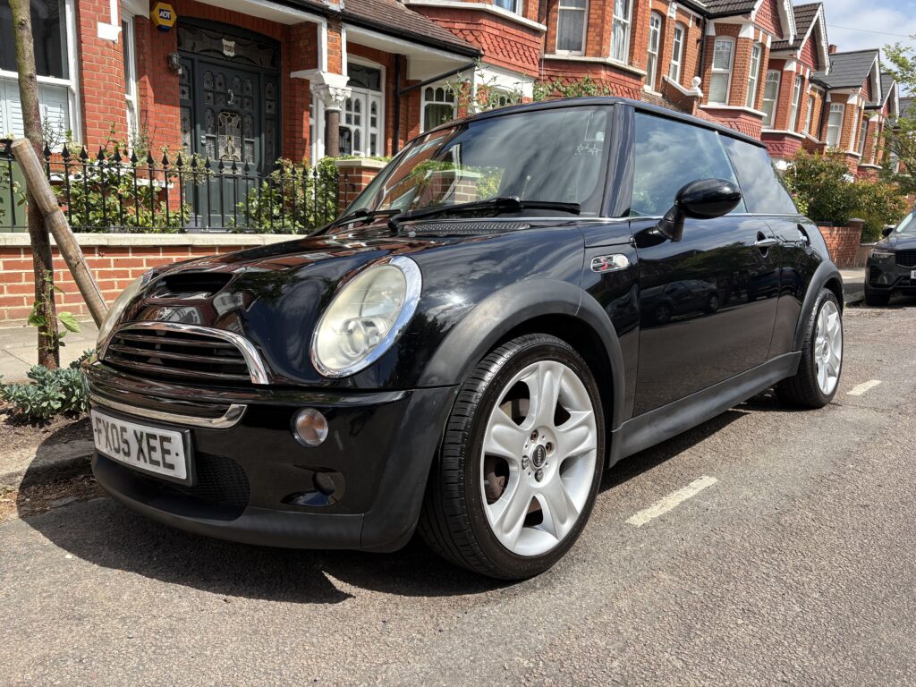 A black Mini Cooper with silver alloy wheels is parked on a residential street. Rows of brick houses with white trim and green doors line the background, while a tree stands on the pavement beside this stylish mini car with UK plate “YX05 XEE.”.