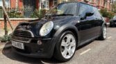 A black Mini Cooper with silver alloy wheels is parked on a residential street. Rows of brick houses with white trim and green doors line the background, while a tree stands on the pavement beside this stylish mini car with UK plate “YX05 XEE.”.