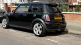 A black mini car with silver wheels is parked on a residential street beside a red brick house with white-trimmed windows and plants in front. The vehicle’s license plate reads “FX05 XEE.” Bright sunlight casts clear shadows on the mini and pavement.