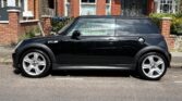 A shiny black Mini car with silver alloy wheels is parked on a residential street in front of red brick houses with white trim and green bushes. The mini is viewed from the side under bright sunlight. Perfect for any Mini trader or enthusiast.